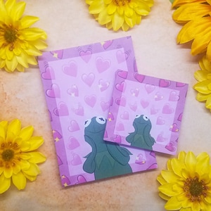 May include: Two pink sticky notes with a green frog character on each. The frog is looking up and has its mouth open. The background of the notes is covered with pink hearts and sparkles.