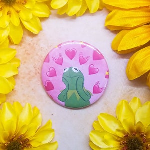 May include: A round button with a pink background and a green Kermit the Frog character. Kermit is looking up with his hands on his cheeks. There are pink hearts with white outlines scattered around him.