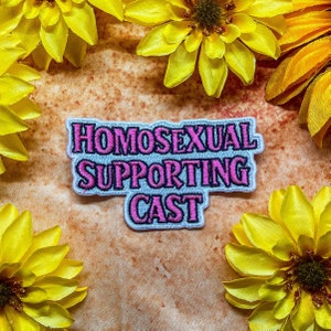 May include: White iron-on patch with pink and black text that reads "HOMOSEXUAL SUPPORTING CAST". The patch is surrounded by yellow flowers.