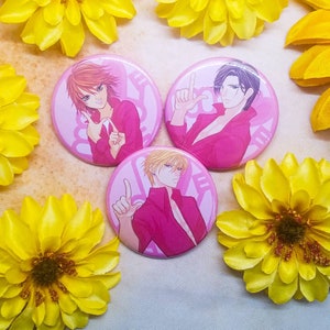 May include: Three pink button pins with anime characters. Each pin features a different character with pink hair, black hair, and blonde hair. The characters are all wearing pink shirts and have a pink background.