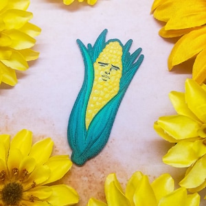 Może przedstawiać: Naszywka z haftowanym kolbem kukurydzy z ludzką twarzą, w kolorze zielonym i żółtym. Kolba kukurydzy jest otoczona żółtymi płatkami kwiatów.