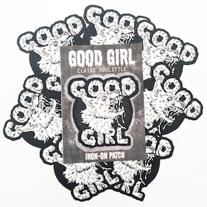 Può includere: Un set di otto toppe termoadesive in bianco e nero con la testa di un lupo circondata dalle parole "GOOD GIRL". Le toppe sono disposte in cerchio con le parole "GOOD GIRL" e "IRON-ON PATCH" visibili su un cartoncino bianco al centro.
