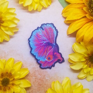 May include: A vibrant pink and blue embroidered patch of a betta fish with flowing fins. The patch is surrounded by yellow flowers.