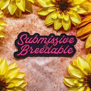Op de afbeelding: Zwarte en roze geborduurde patch met de tekst "Submissive & Breedable" op een bruine achtergrond met gele bloemen.