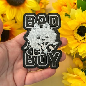 May include: A black embroidered patch featuring a white fluffy dog wearing a spiked collar. The text BAD BOY is stitched in white block letters above and below the dog. The patch has a black border and a detailed white thread design.