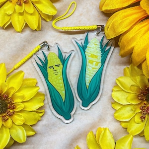 Puede incluir: Dos llaveros coloridos con maíz en la mazorca de dibujos animados con caras. El maíz es amarillo y verde sobre un fondo blanco. Los llaveros están unidos a una cuerda blanca.