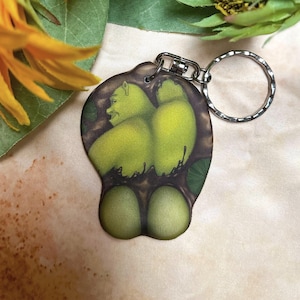 May include: A green and brown key chain featuring a cartoon character with large green eyes and a wide smile. The character is depicted in a sitting position with its legs crossed. The key chain is attached to a silver metal ring.