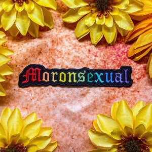 May include: A black embroidered patch with the word "Moronsexual" in rainbow colours. The patch is surrounded by yellow flowers.