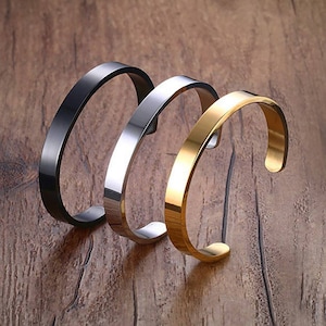 Op de afbeelding: Drie metalen armbanden in verschillende afwerkingen: zwart, zilver en goud. De armbanden zijn eenvoudig en stijlvol, met een afgeronde vorm.