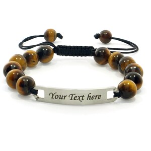 Op de afbeelding: Een bruin en gouden tijgeroog-parel armband met een zwart koord en een zilveren tag met de tekst "Your Text here".