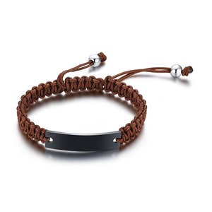 May include: A brown braided bracelet with a black metal nameplate. The bracelet has a sliding knot closure and two silver beads.