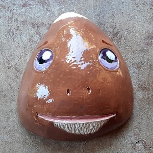 May include: A brown ceramic sculpture of a face with white and purple details. The face has a wide smile and two large eyes.