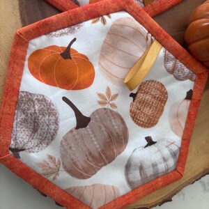 May include: A hexagonal fabric potholder featuring a white background with a repeating pattern of orange, brown, and beige pumpkins and small leaves. The edges are finished with an orange fabric binding and a small yellow hanging loop is attached.