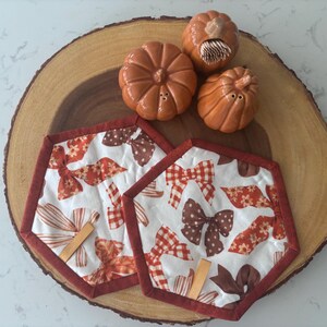 May include: Two hexagonal fabric pot holders featuring a white background with a repeating pattern of brown and orange bows, finished with a dark red border and a small tan tab. Three ceramic pumpkin-shaped shakers in shades of orange.