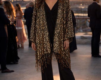 May include: A woman wearing a black jumpsuit and a gold sequined jacket with fringe. The jacket is open, revealing the jumpsuit underneath. The background shows a sunset and people at an outdoor event.