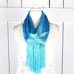 Peut inclure: Une écharpe turquoise à franges avec une finition chatoyante. L'écharpe est drapé autour d'un haut en dentelle blanche sur un mannequin.