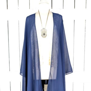 May include: A navy blue chiffon kimono-style wrap on a white mannequin. The open-front design features long, flowing panels. A silver-toned pendant necklace with a large, ornate charm is visible. The background is a white, vertical-slat wall.