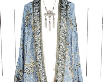 Puede incluir: Una chaqueta estilo kimono con estampado de cachemira en azul claro y diseño drapeado. La chaqueta presenta un patrón detallado en tonos grises y dorados. Se ve un collar plateado con un colgante.