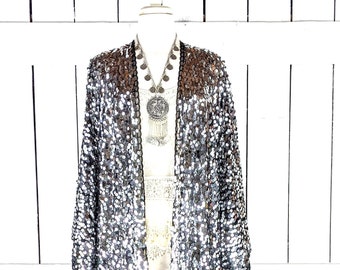 May include: A long, open-front sequin kimono in silver and black, displayed on a mannequin. The kimono has a fringe hem and is paired with a white top and a silver necklace with a large pendant. The background is a white wooden wall.