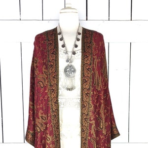 May include: A long, burgundy paisley patterned kimono with gold accents and fringe. The kimono is draped over a white dress form.