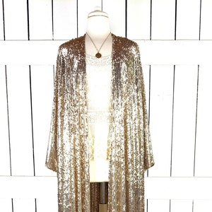 May include: A long, gold sequined cardigan with a shawl collar and long sleeves. The cardigan is open in the front and is being displayed on a white mannequin.