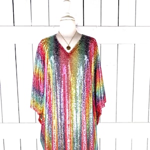 May include: A multi-coloured sequin kaftan dress with a V-neck and wide sleeves. The dress is displayed on a mannequin and features vertical stripes of various colours, including red, yellow, green, and blue. A gold-coloured necklace is visible.