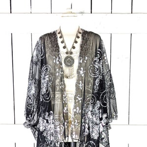 May include: Black sheer kimono with white floral embroidery and fringe trim. The open-front design features a flowing silhouette. A silver necklace with a large pendant and coin accents is visible.