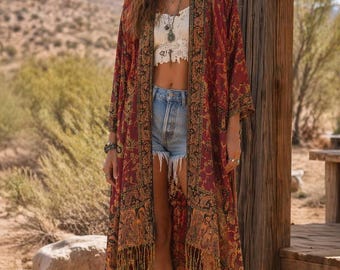 Puede incluir: Un kimono largo y fluido de color burdeos con un estampado de cachemira y ribete de flecos. El kimono se lleva con un top corto de encaje blanco, pantalones cortos de mezclilla y botas de vaquero marrones. El fondo es un paisaje desértico.