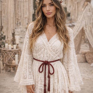May include: A cream-colored lace dress with long, flared sleeves and a V-neckline. The dress is cinched at the waist with a burgundy rope belt. The model is wearing a floral crown and layered gold necklaces.
