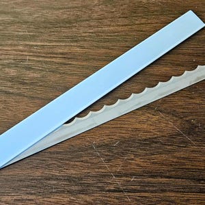 May include: Two cake decorating tools are shown on a wooden surface. One is a light blue plastic guide, and the other is a serrated metal blade. The blade has a hole at one end. The tools are designed for cake decorating.