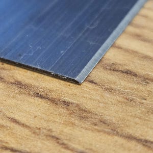 May include: Close-up of a metal blade with a sharp edge, set against a wooden surface. The blade has a dark, reflective surface, and the wood grain is visible in the background. The blade's edge is angled.
