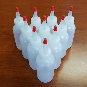 May include: Ten clear plastic bottles with red caps and a pointed tip. The bottles are arranged in a triangle shape on a wooden surface.