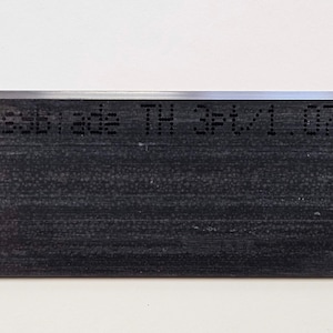 Op de afbeelding: Close-up van een zwart metalen blad met de tekst "Essabrade TH 3#tv1.07 0.997" gegraveerd in het oppervlak.