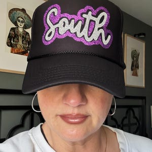 May include: Black trucker hat with the word "South" in a purple and silver glitter design. The hat has a curved brim and a mesh back. The hat is worn by a person.