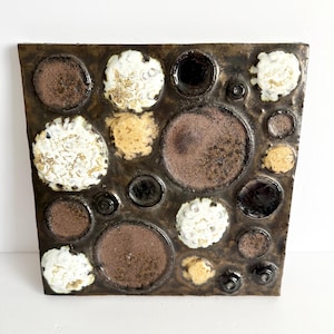 May include: Square ceramic tile with a dark brown background and a pattern of various-sized circles. The circles are in shades of brown, white, yellow, and black, with a textured, glazed finish. The tile has a vintage, handcrafted aesthetic.