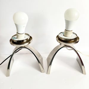 May include: Two modern, silver-toned table lamps. Each lamp features a curved base and a circular top holding a white LED light bulb. The lamps are designed with a minimalist aesthetic, suitable for contemporary decor.