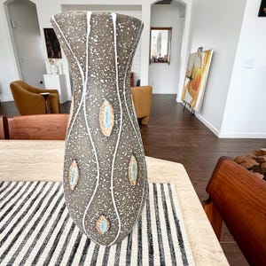 May include: A tall, tapered ceramic vase with a textured dark gray surface featuring white speckled paint splatters. The vase is decorated with vertical white wavy lines and several raised, almond-shaped accents in light tan with blue centers.