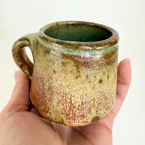May include: A small, handmade ceramic mug with a brown, green, and yellow glaze. The mug has a rounded body and a small handle.