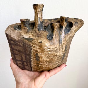 May include: A hand-built ceramic vessel with a textured, uneven surface featuring a mix of tan, brown, and black glazes. The object has four distinct, chimney-like openings at the top and a dark brown grid pattern etched onto one side of the exterior.