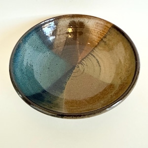 May include: A handmade ceramic bowl with a unique color scheme. The bowl is divided into four sections, each with a different color: teal, brown, tan, and a darker brown. The bowl has a glossy finish and a circular shape.