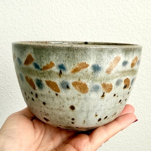 May include: A ceramic bowl with a speckled light gray base, featuring a horizontal band of alternating brown and blue brushstroke patterns. The lower portion of the bowl is decorated with small dark brown speckles, and the rim has a muted green tint.