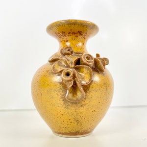 May include: A small, yellow ceramic vase with a textured surface and brown floral accents. The vase has a round body and a narrow neck.