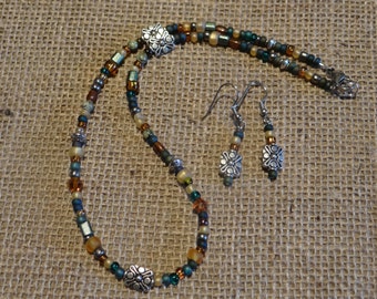 Könnte beinhalten: Ein Halsketten- und Ohrring-Set mit braunen, grünen und silbernen Perlen. Die Halskette hat einen silbernen Blumenanhänger und die Ohrringe haben silberne Blumenanhänger.