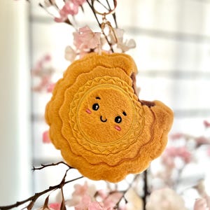 May include: A plush keychain shaped like a mooncake with a smiling face. The keychain is yellow and has a brown bite taken out of it. The keychain is hanging from a branch of a pink flowering tree.