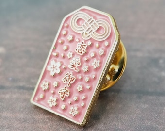 May include: A pink enamel pin with a gold border and a white knot design. The pin features Japanese characters and a floral pattern.