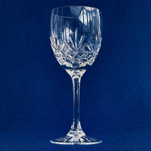 Può includere: Un bicchiere da vino di cristallo trasparente con un lungo stelo e un design dettagliato intagliato sulla coppa. Il bicchiere è posto su uno sfondo blu scuro, evidenziando i suoi intricati motivi e la sua forma elegante.