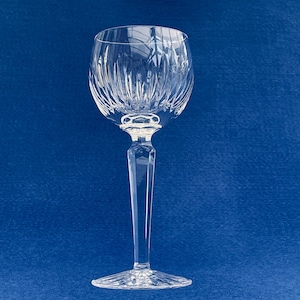 May include: A clear crystal wine glass with a long stem and a detailed, textured bowl. The glass is set against a solid blue background. The bowl has vertical cuts, and the stem is faceted. The base is round and also textured.