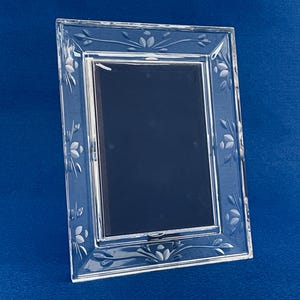 Puede incluir: Un marco de fotos rectangular de vidrio transparente con diseños florales y de hojas grabados. El marco tiene un borde biselado y un fondo azul oscuro. El marco está ligeramente inclinado, mostrando sus detalles decorativos.