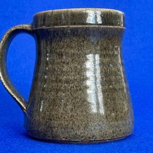 Puede incluir: Una taza de cerámica gris con una asa ancha. La taza tiene un esmalte moteado y un labio ligeramente acampanado.