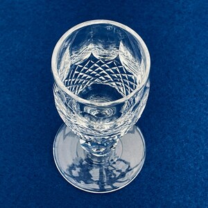 May include: A clear, cut crystal wine glass with a round base and a diamond pattern on the bowl. The glass is set against a deep blue background, showcasing its intricate design and clarity.